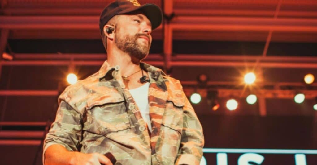 Chris Lane stands on stage in a camouflage shirt and hat, delivering an energetic performance to an enthusiastic crowd.