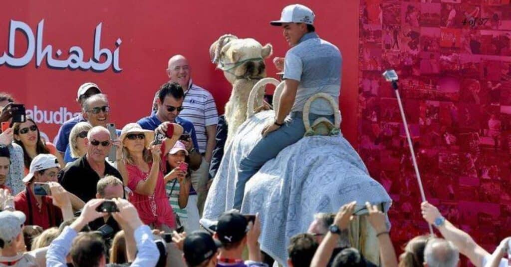 Rickie Fowler rides a camel, captivating a crowd with his adventurous spirit in this vibrant outdoor setting.