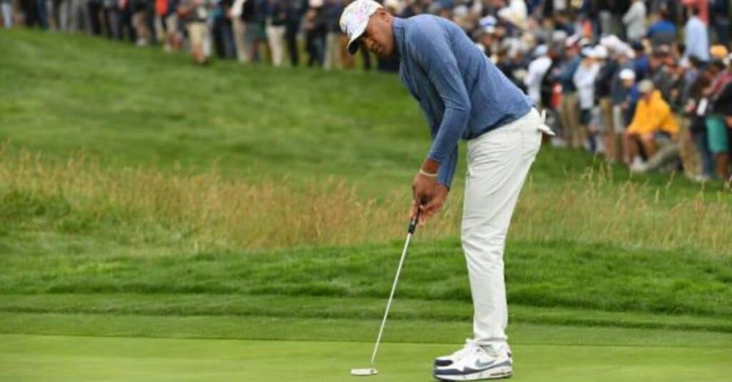 Tony Finau, dressed in white pants and a blue shirt, is poised to putt a golf ball on the course.