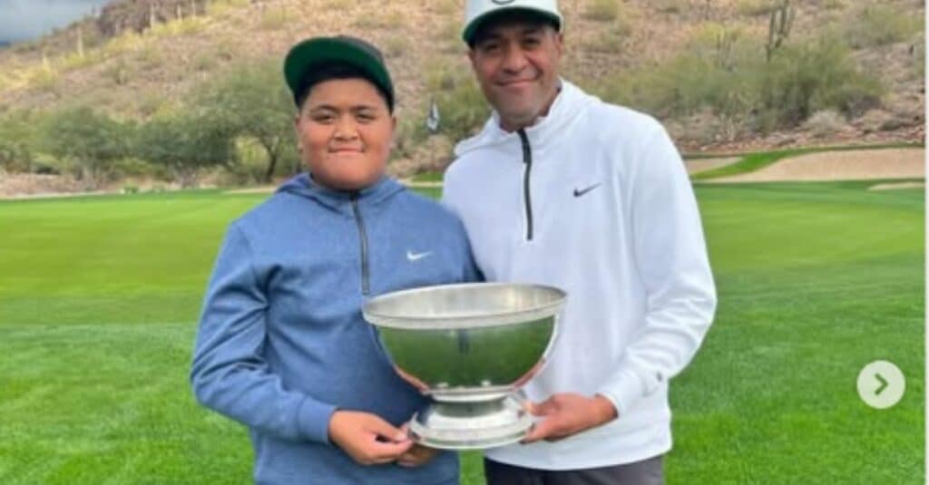 Tony Finau and a companion celebrate their success by holding a trophy on a beautiful golf course backdrop.