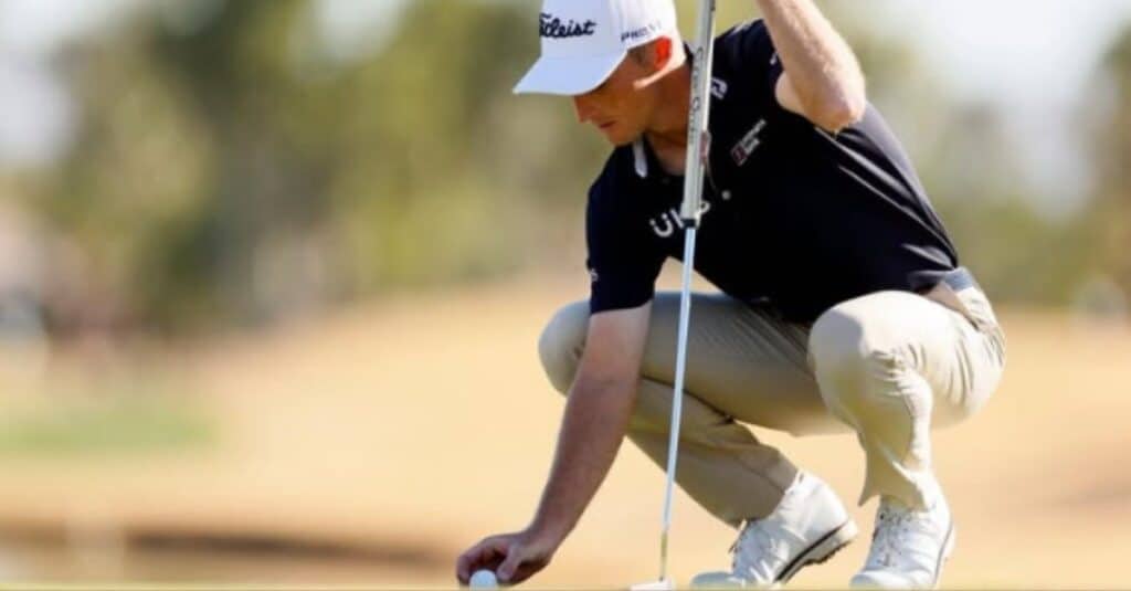 Will Zalatoris kneels in a black shirt and white hat, preparing to place a golf ball on the green with concentration.