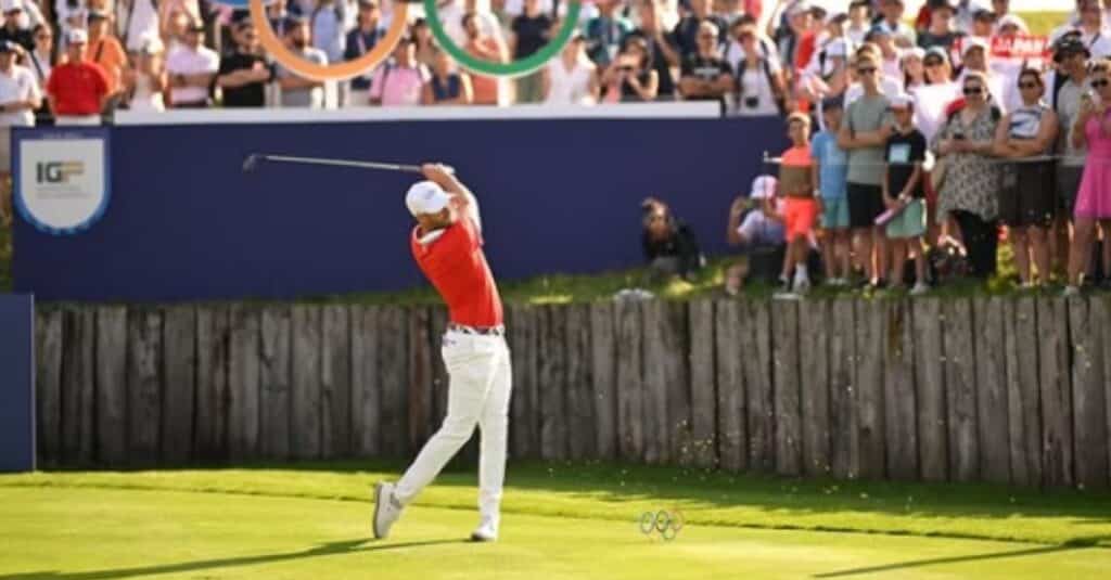 Wyndham Clark, dressed in a red shirt, swings a golf club as a crowd watches intently.