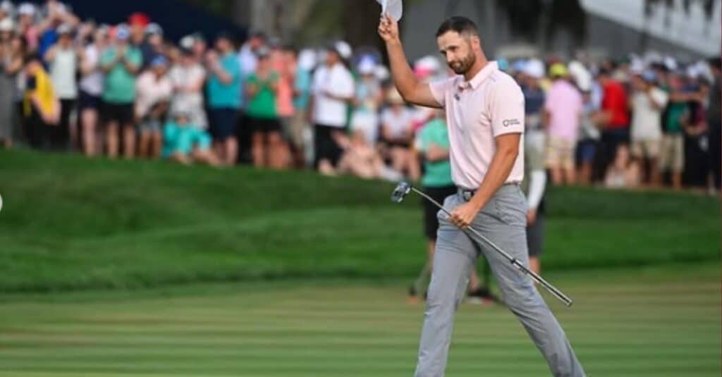 Wyndham Clark, dressed in a pink shirt, grips a golf club, poised for action on the golf course.