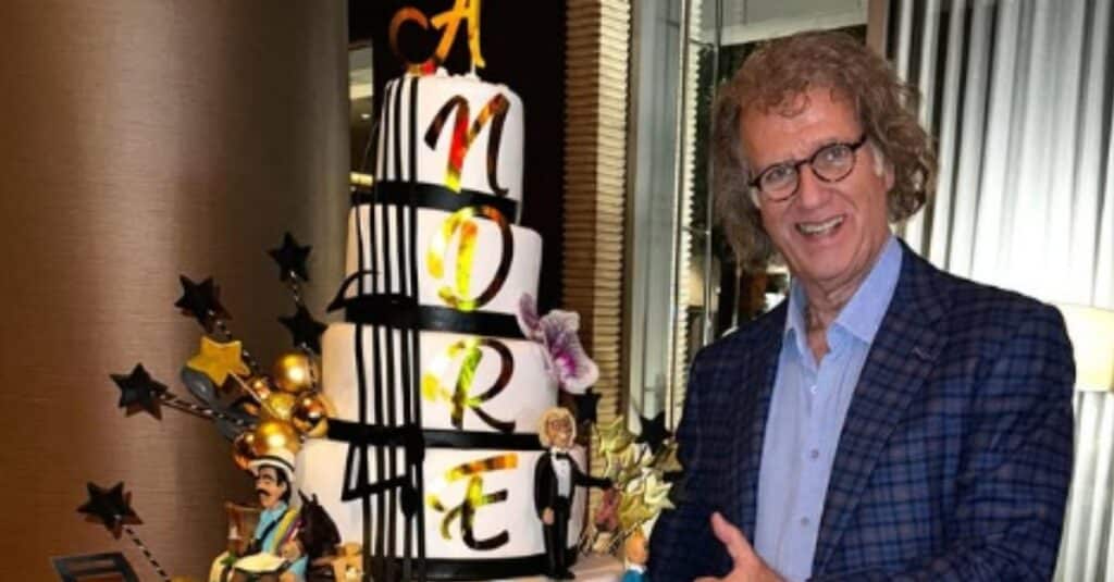 Andre Rieu, wearing glasses, stands next to an elegantly designed cake, showcasing a joyful moment.