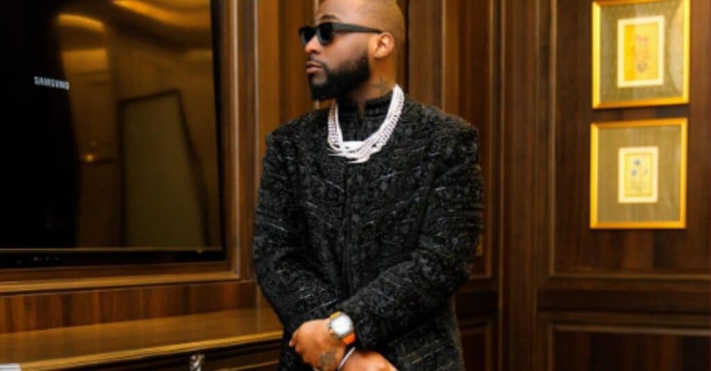 Davido, dressed in a black suit and sunglasses, stands poised in front of a television, exuding charisma and elegance.