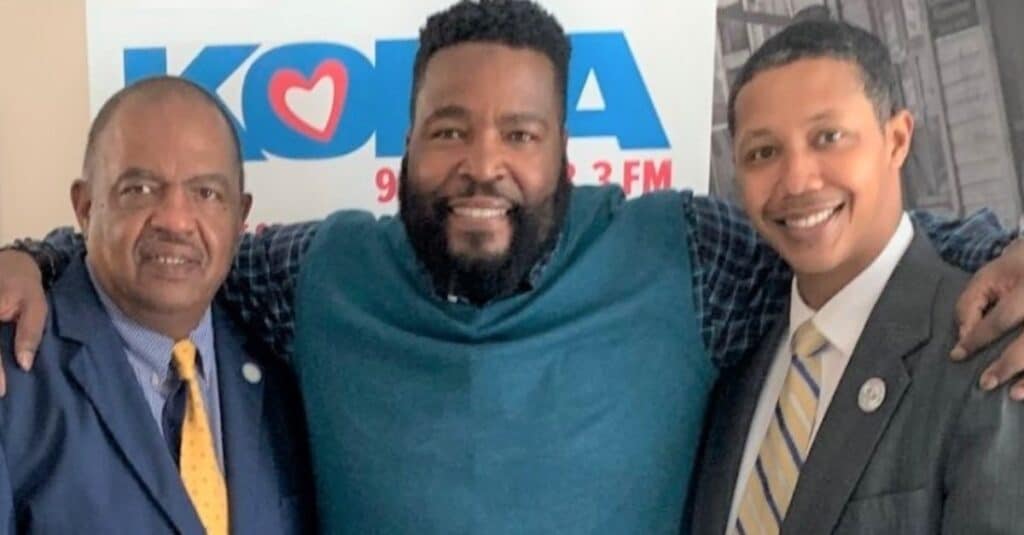 Dr. Umar Johnson stands with two friends, posing cheerfully in front of a koa radio sign.