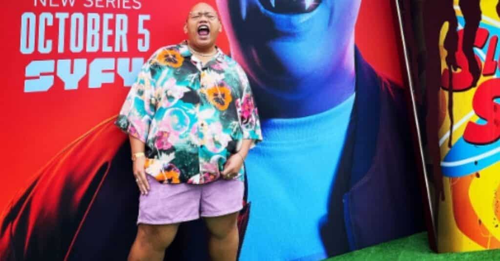 Jacob Batalon, dressed in shorts and a floral shirt, poses in front of a vibrant poster.