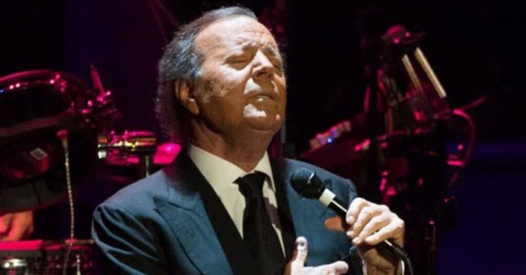 Julio Iglesias, dressed in a suit, passionately sings into a microphone during a live performance.