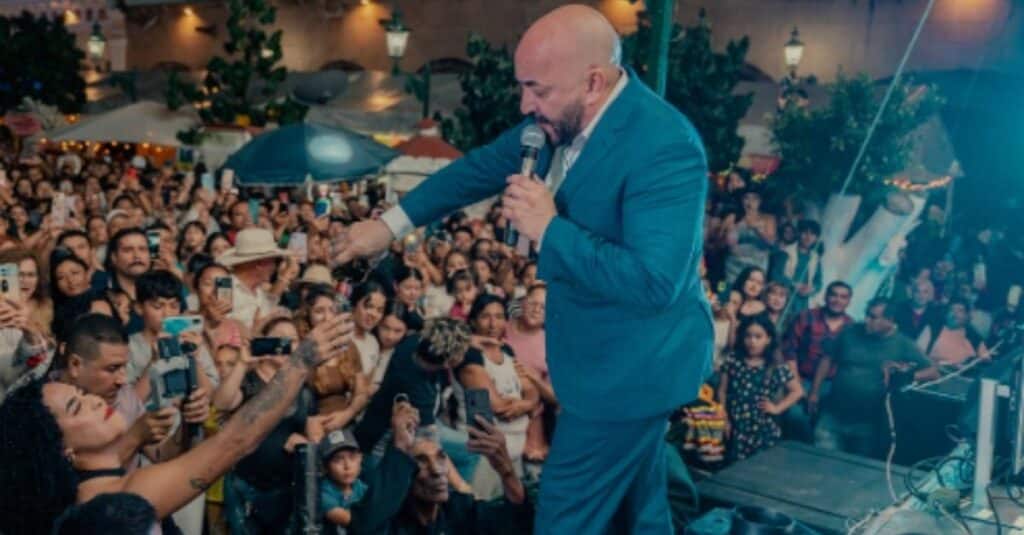 Lupillo Rivera, dressed in a suit, stands on stage with a microphone, poised to engage with the audience.