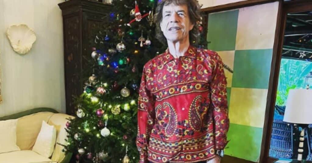 Mick Jagger stands in front of a beautifully decorated Christmas tree, exuding holiday spirit and charm.