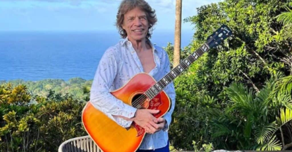 Mick Jagger stands with an orange guitar, framed by a picturesque view of the ocean.